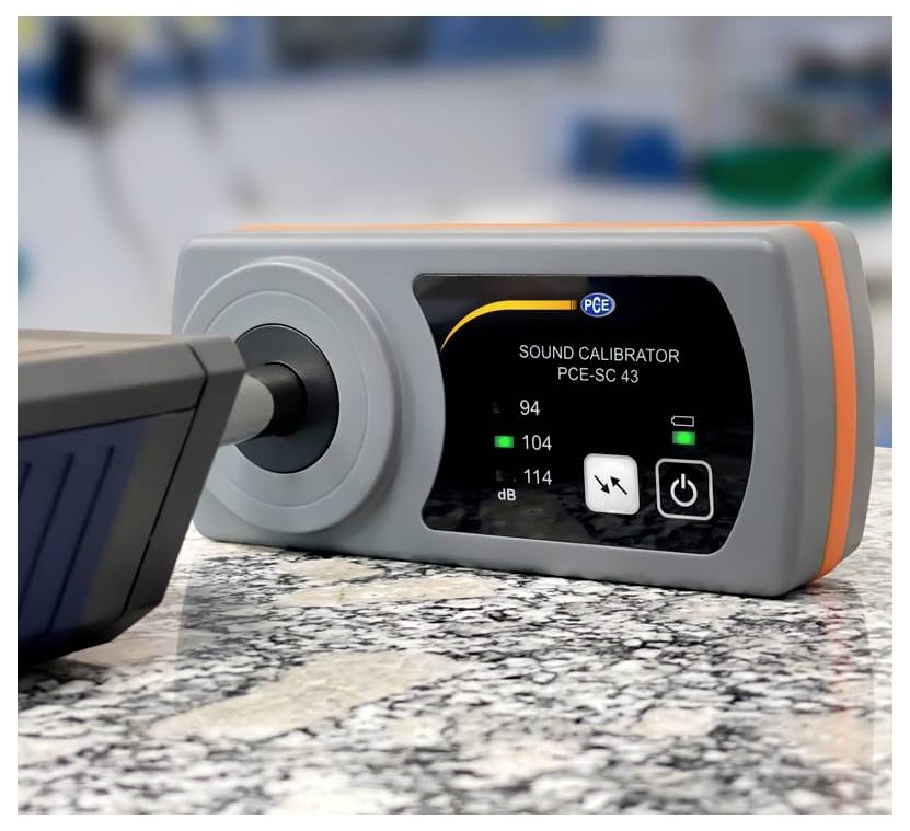 Geluidskalibrator op een werkblad, met een luidspreker of microfoon in de voorkant en een digitaal scherm dat geluidsniveaus en stromaansluitingen toont.