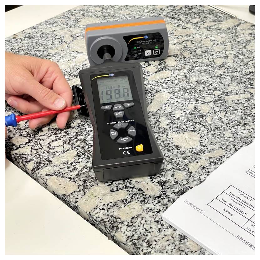 Hand die een geluidsniveaumeter tegen een granieten aanrechtblad houdt, terwijl een ander meetinstrument in de buurt staat en oppervlaktegeluid of trillingen meet.