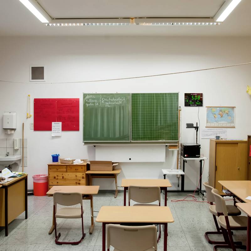 Leeg klaslokaal met schoolbord, tafels en stoelen. Een wereldkaart en klok aan de muur.