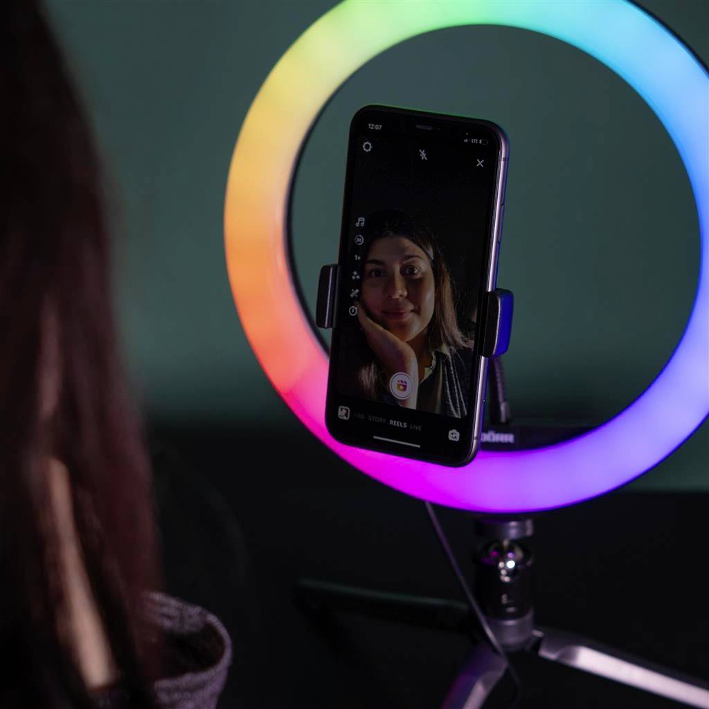 Een persoon maakt een selfie met een smartphone die is bevestigd in een gekleurde ringlichthouder, in een donkere ruimte.