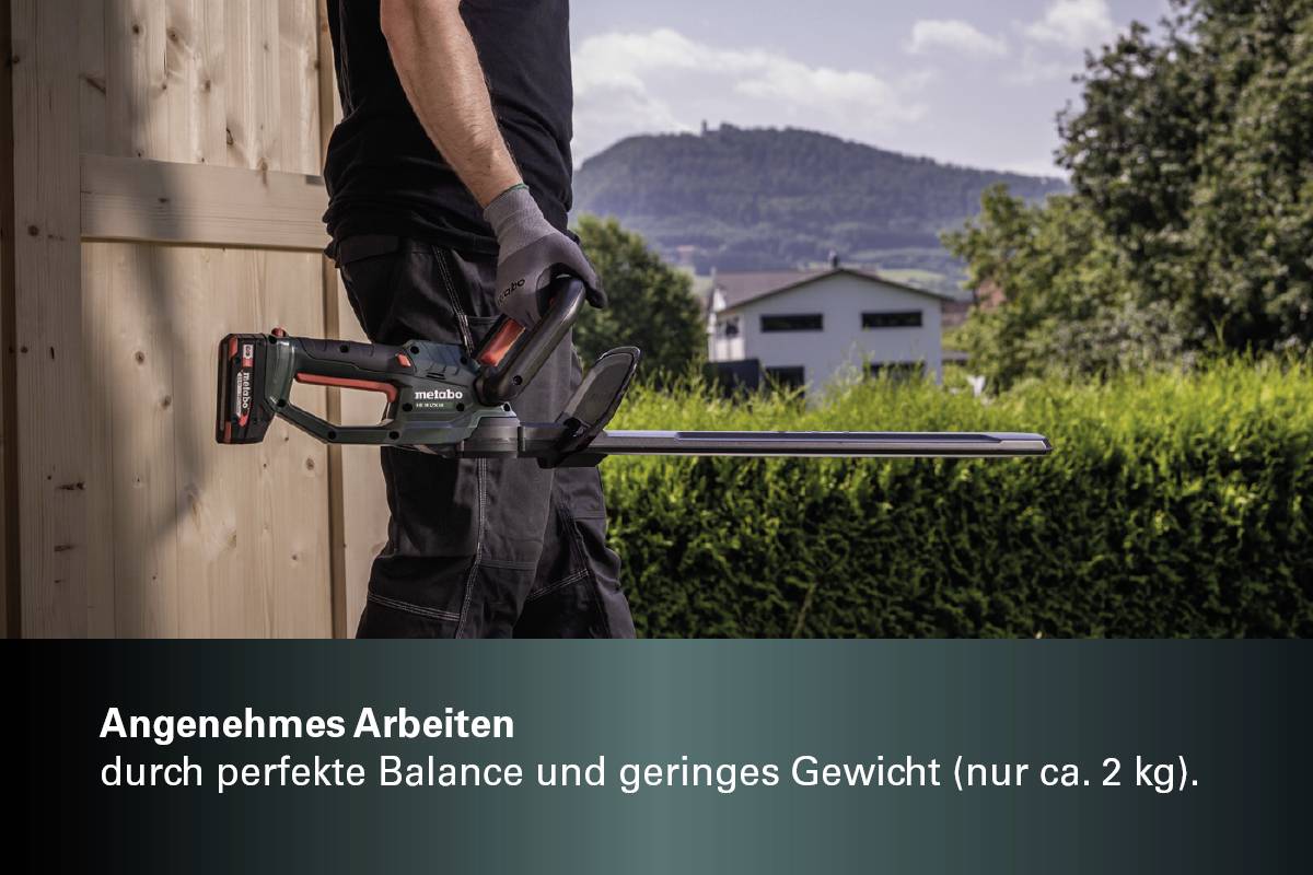 Een persoon snoeit hagen met een elektrische heggenschaar. Op de achtergrond zijn een houten schuur, struiken en een bebost heuvellandschap zichtbaar.