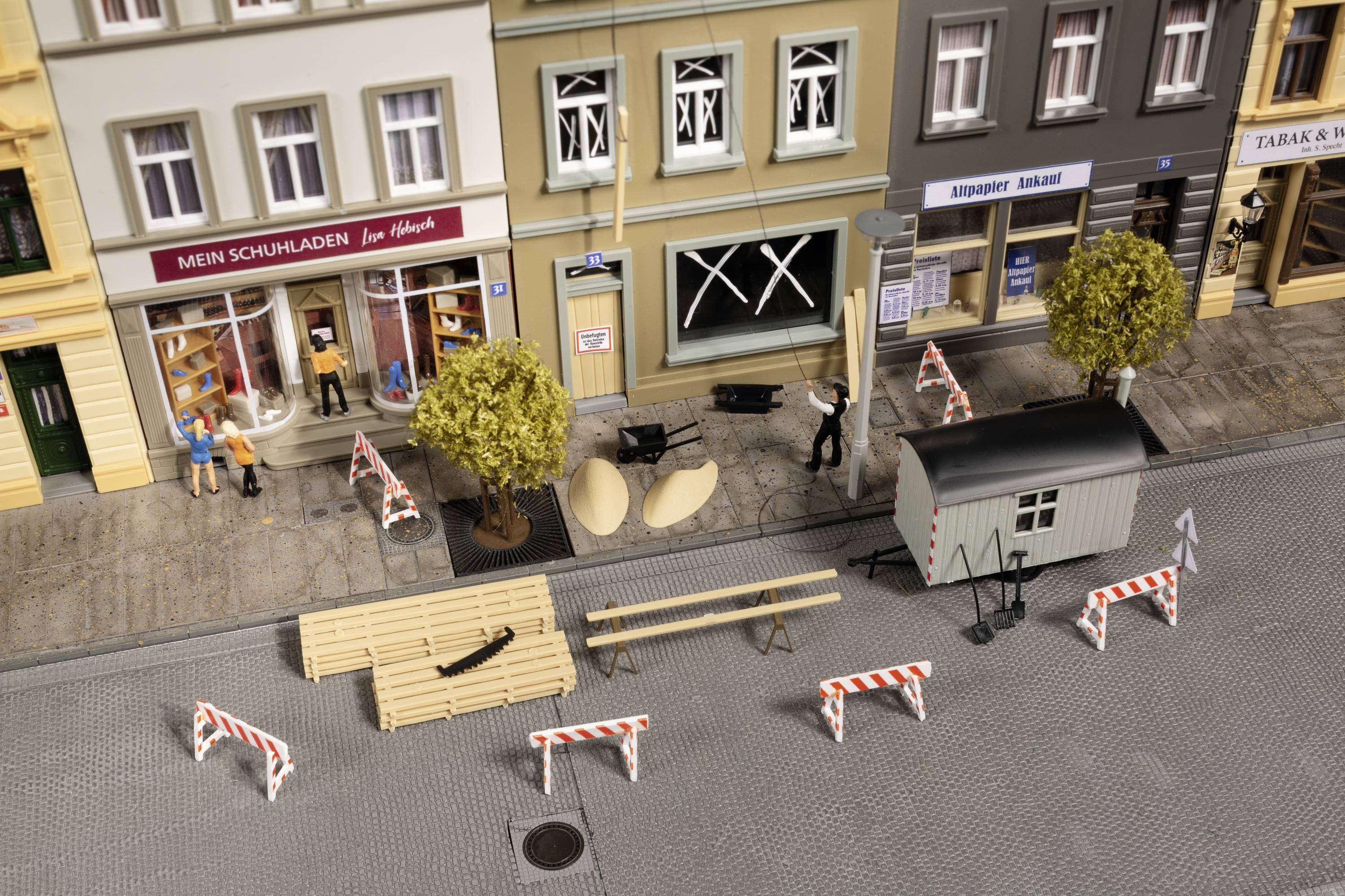 Straatscène met miniatuur-bouwplaats: afzettingen, stapels hout en containers op het voetpad. Gebouwen op de achtergrond deels beklad.