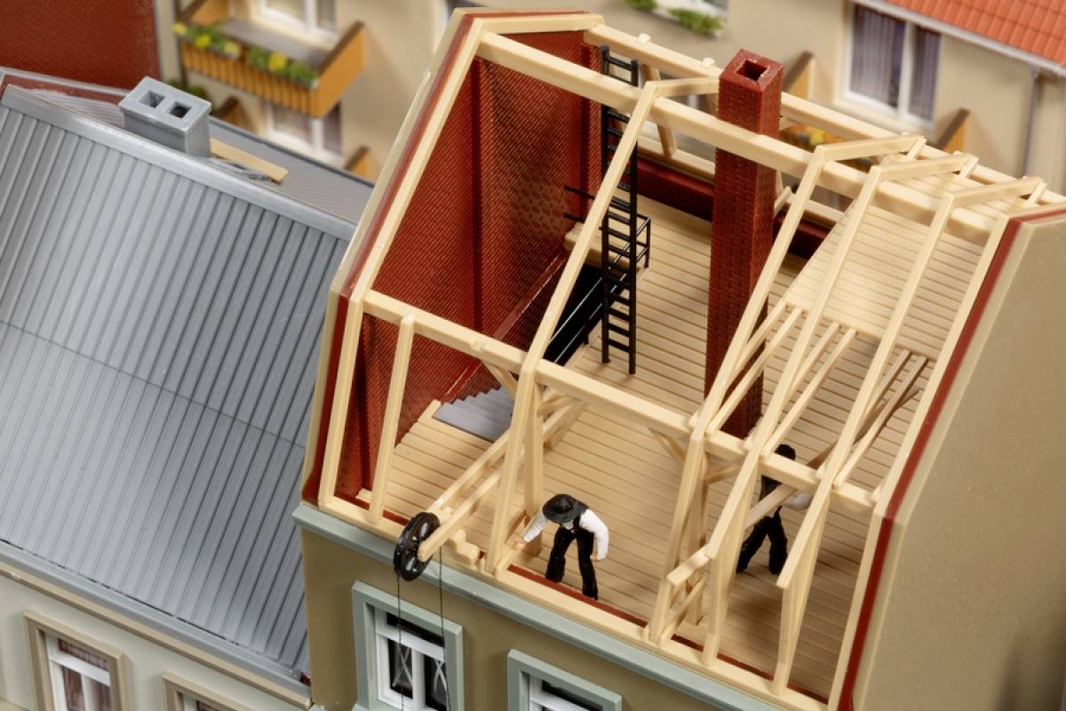 Een modelhuis met gedeeltelijk open dak toont een houten dakconstructie. Twee figuren werken aan de bouw van het dak.