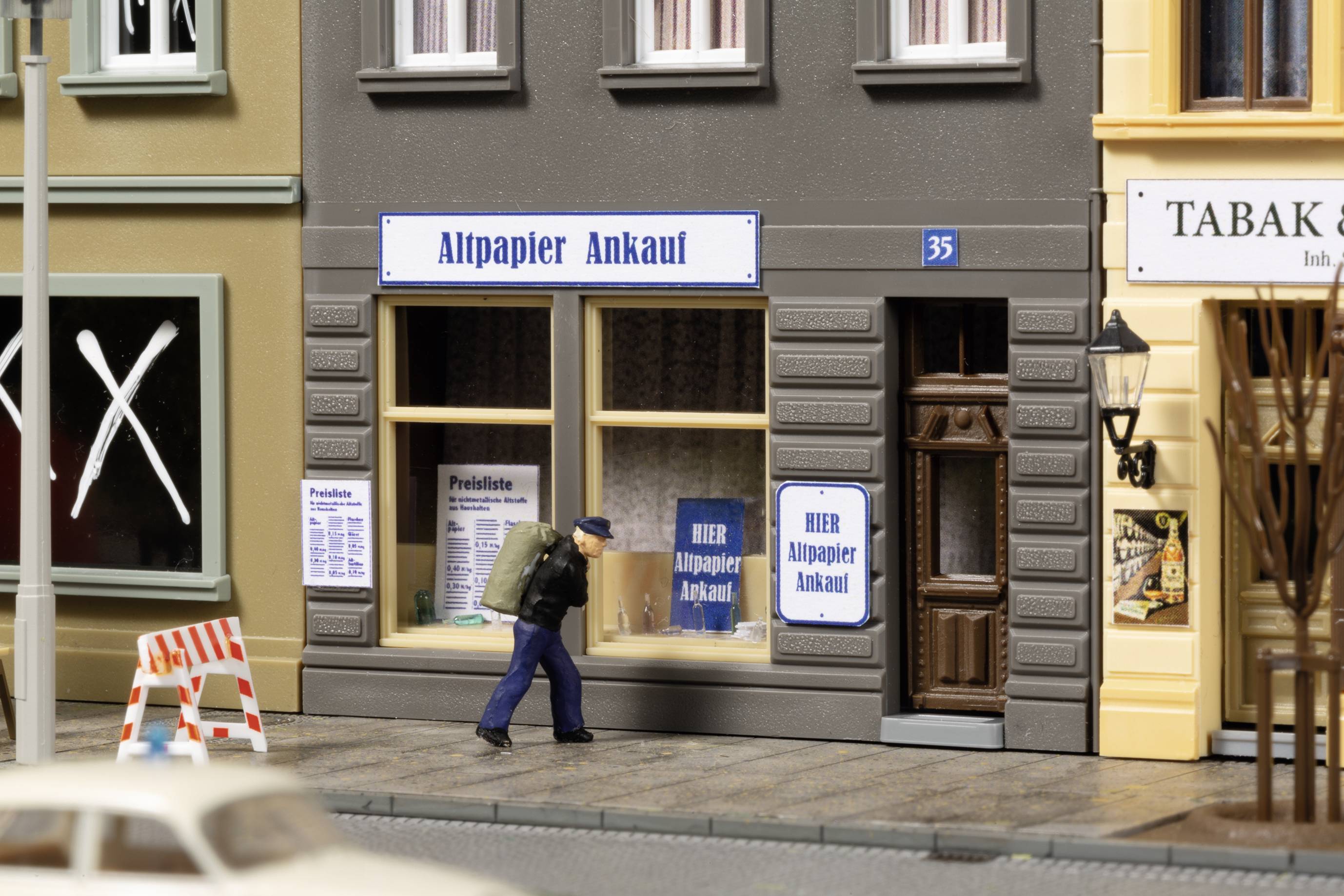 Een miniatuurstadsscène toont een man die over het voetpad langs een winkel voor oud papieraankoop loopt.