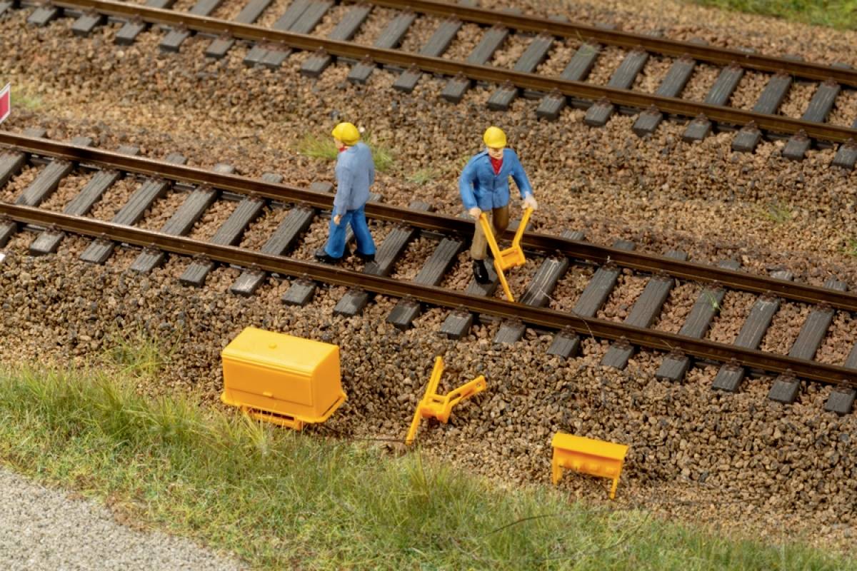 Modelspoorbaan-scène met twee figuren in werkkleding op sporen, die spoorgereedschap gebruiken. Omringd door kleine gereedschapskisten.