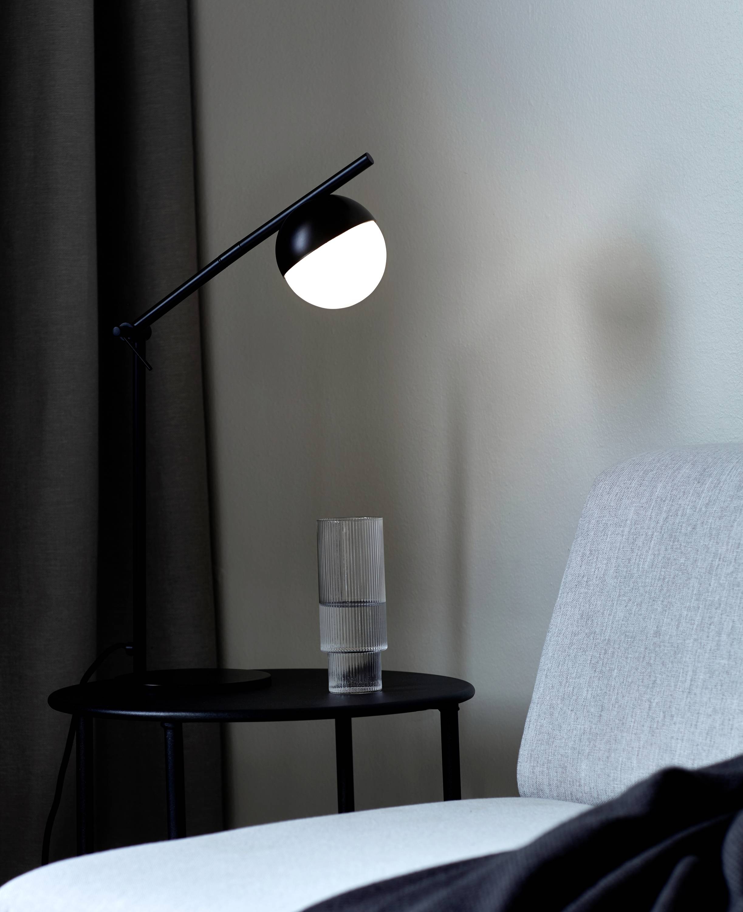 Een moderne lamp met een ronde lichtbron staat op een rond bijzettafel naast een grijze bank, met een glas water ernaast.