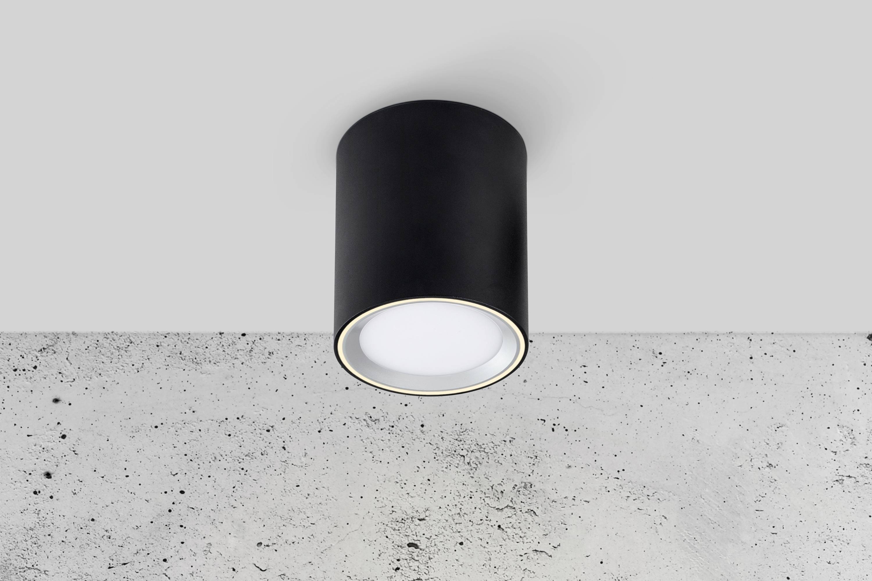 Een cilindrische, zwarte plafondlamp met wit licht is gemonteerd aan een grijs gemarmerd plafond.