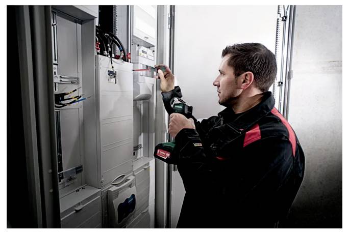 Een technicus in een zwart en rood uniform gebruikt een boormachine om te werken aan een elektrisch paneel in een technische ruimte, wat onderhoudswerkzaamheden aangeeft.