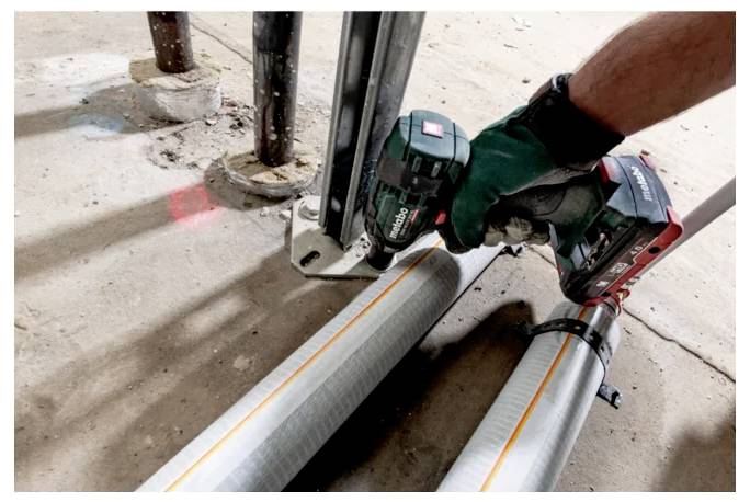 Een persoon met handschoenen gebruikt een elektrisch gereedschap om metalen buizen op de vloer te bevestigen. De buizen hebben gele lijnen en zijn aan elkaar geklemd.
