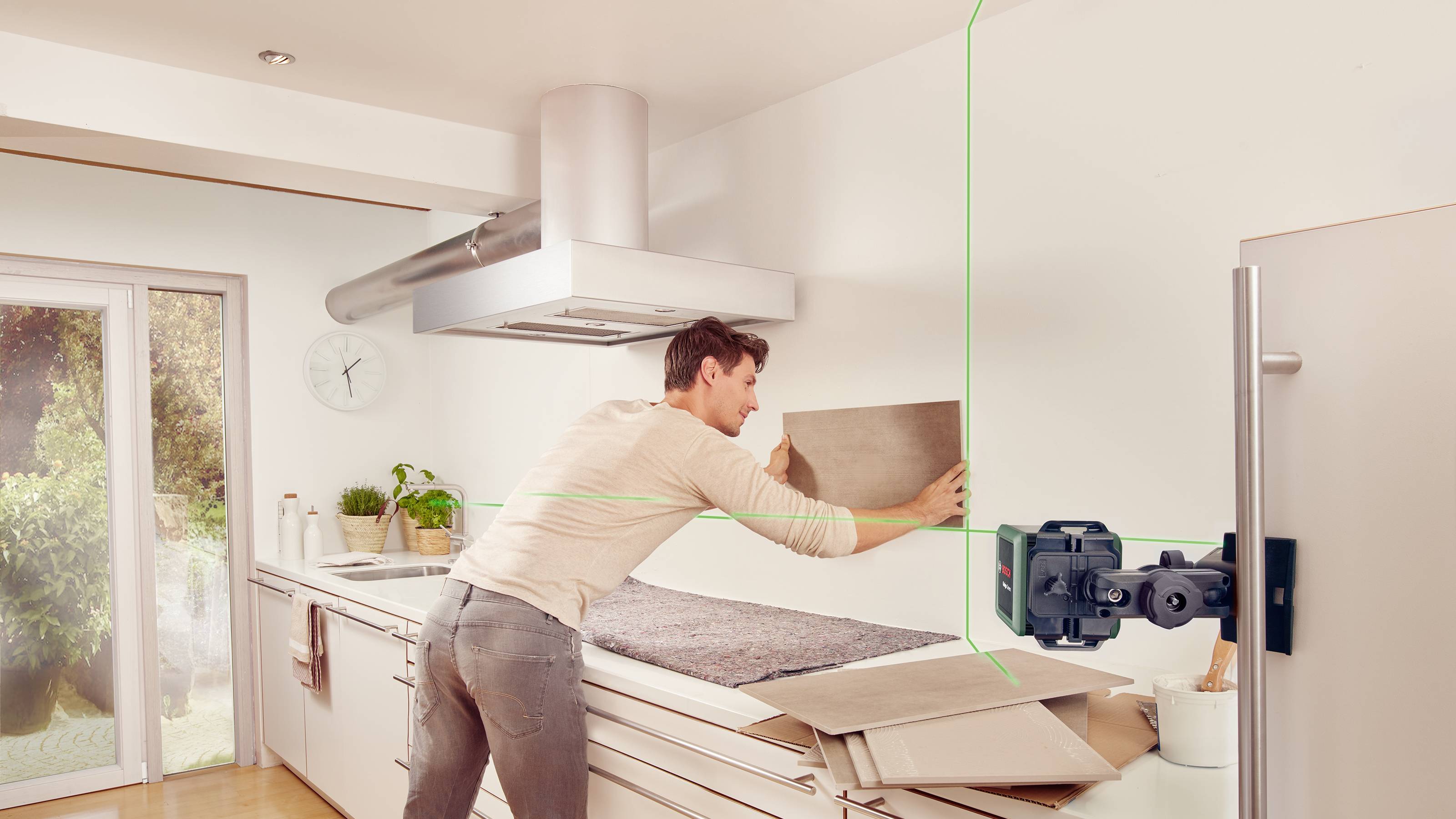 Een man richt een tegel aan een keukenmuur met behulp van een laser-waterpas. Op de achtergrond een moderne keuken met daglicht.