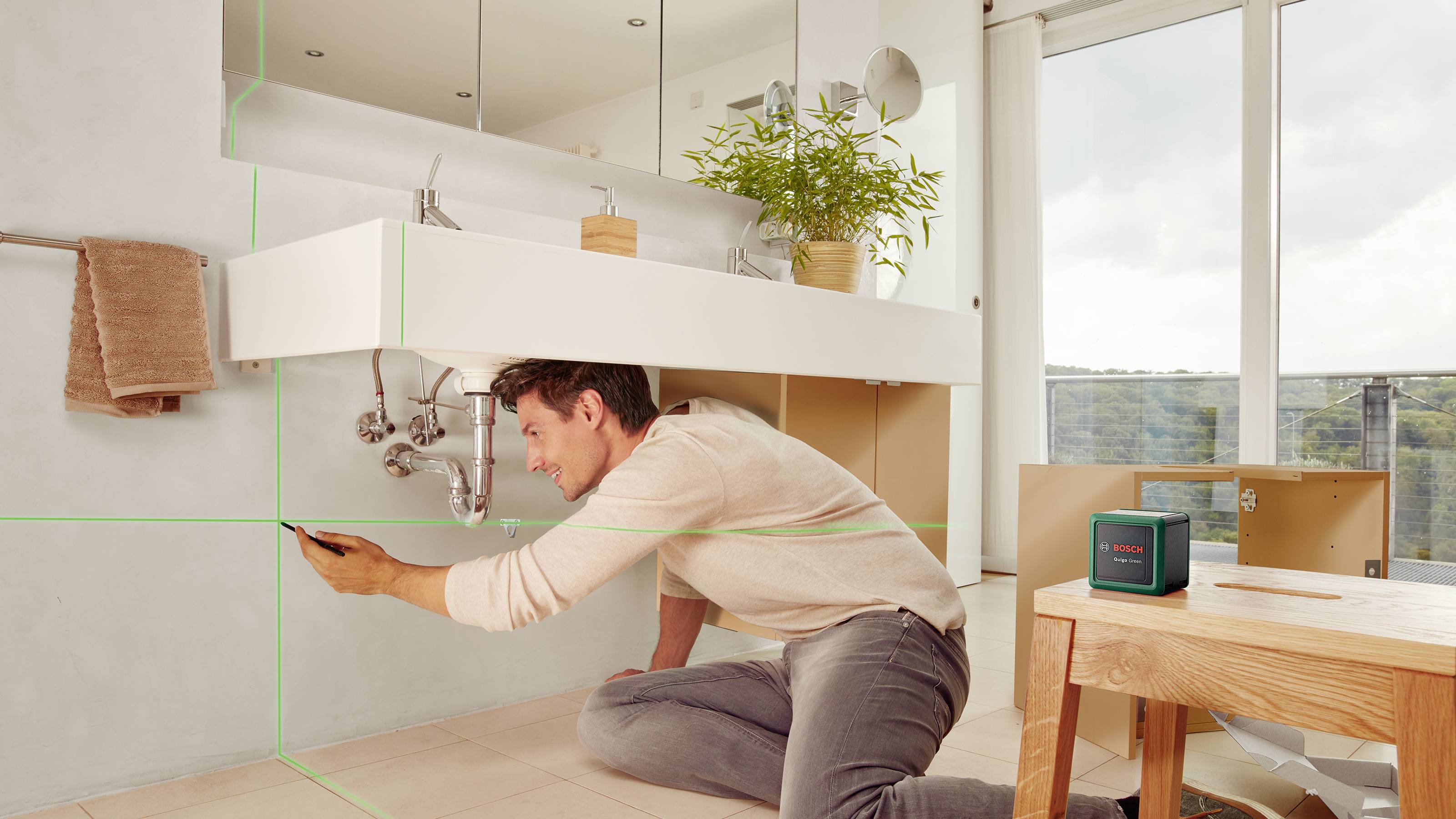 Een man richt een laser voor hoogtemeting in een badkamer. Achter hem bevindt zich een raam met zicht op bomen.
