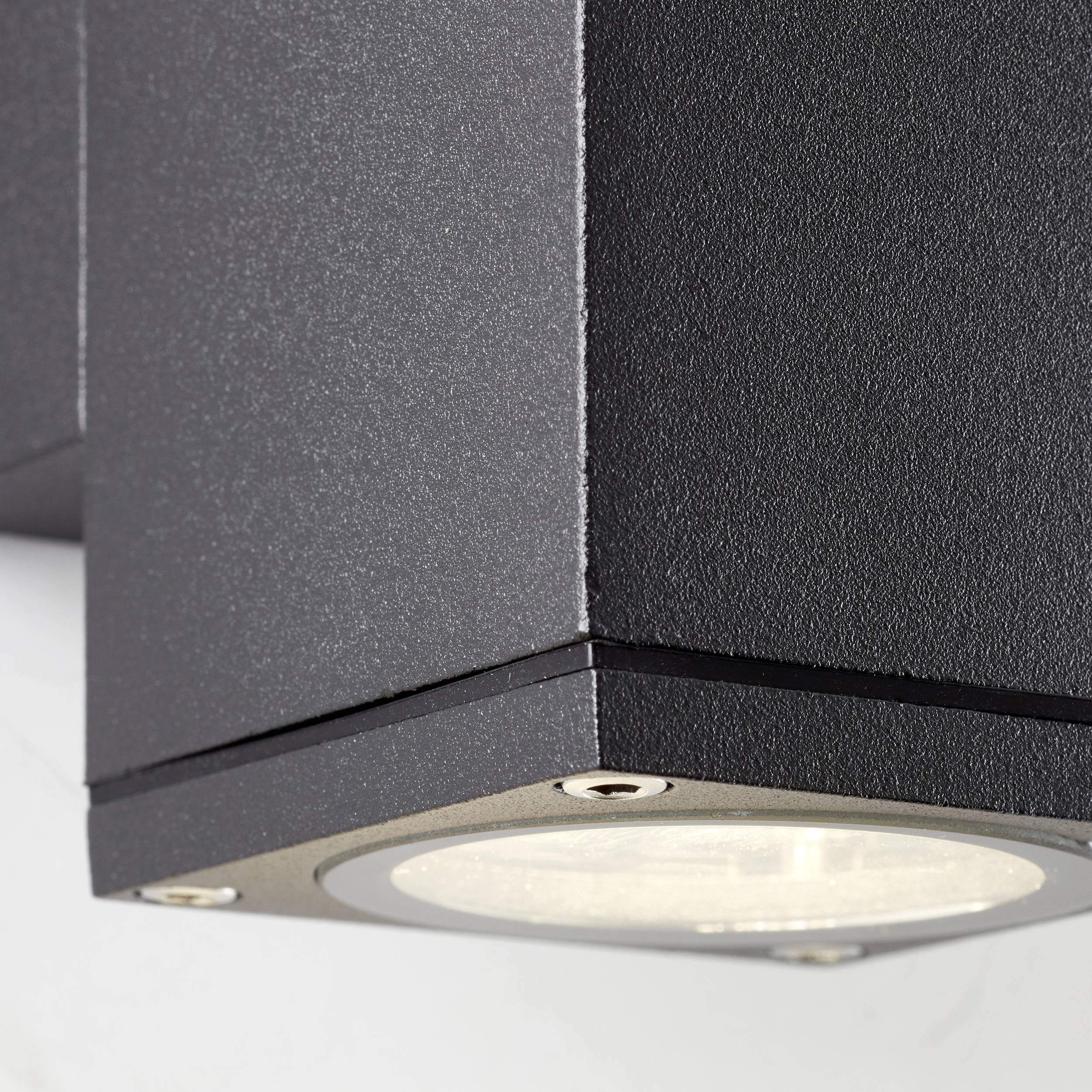 Zwarte, zeshoekige buitenwandlamp met heldere glazen afdekking, gemonteerd op een witte oppervlakte.
