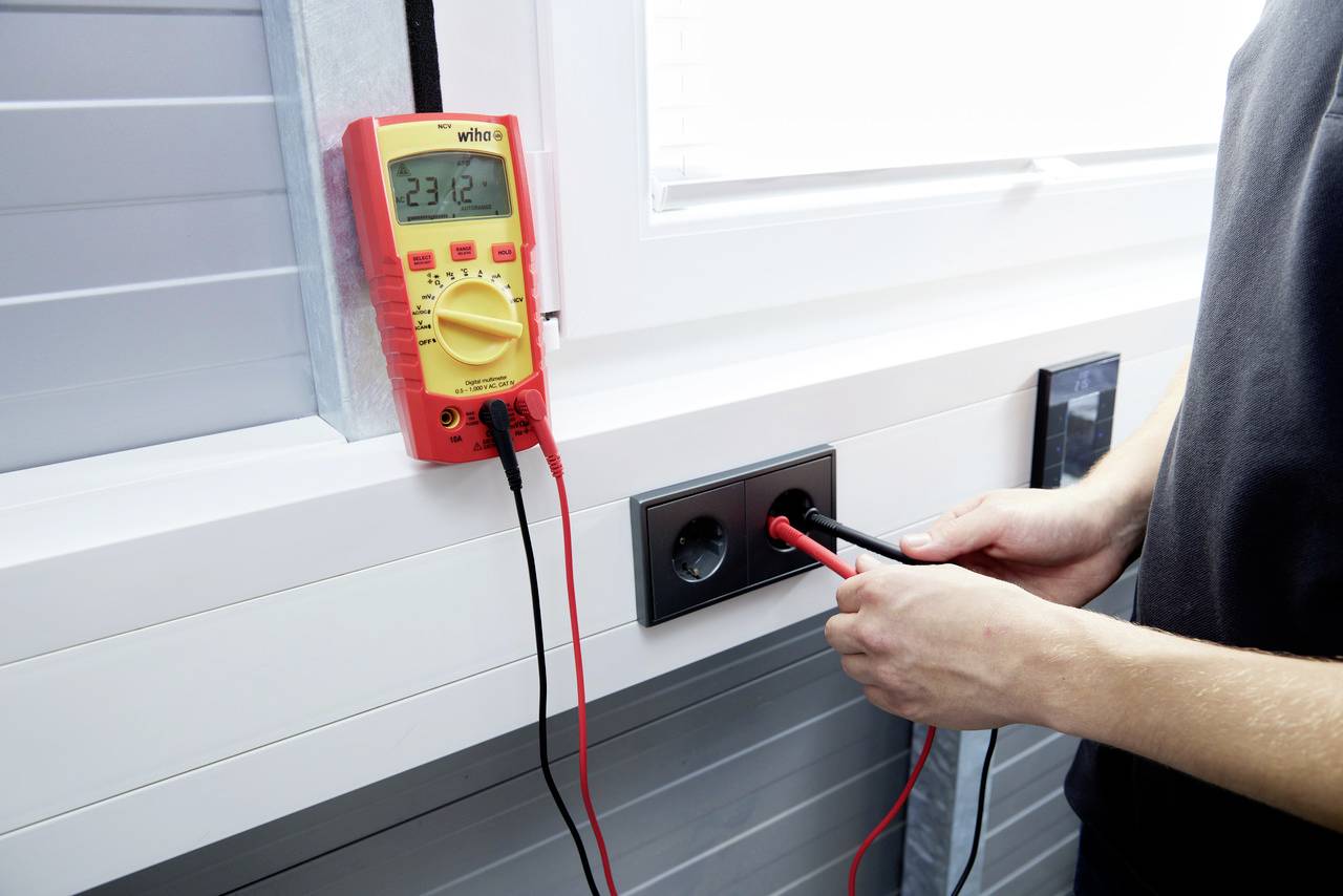Een persoon meet de spanning bij een stopcontact met een multimeter. Het display toont '23.12'. De scène speelt zich af bij een vensterraam.