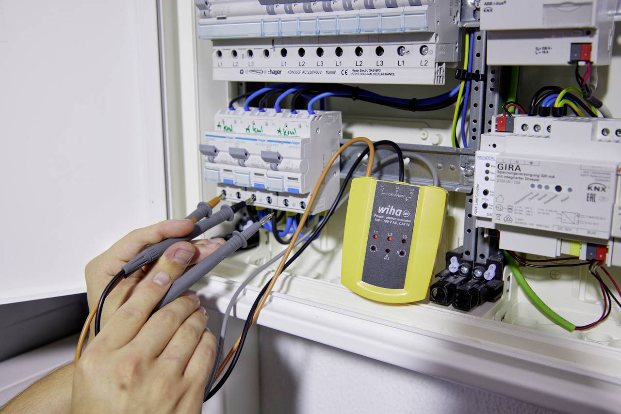 Een persoon meet stroomwaarden met meetapparatuur aan een open elektriciteitskast. Verschillende kabels zijn aangesloten, meetapparaat toont waarden.