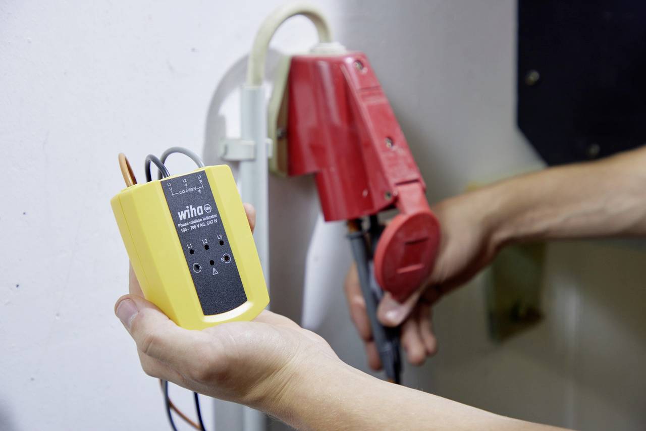 Een persoon houdt een geel elektrisch meetapparaat naast een rode stekkerverbinding in de buurt van een muur.