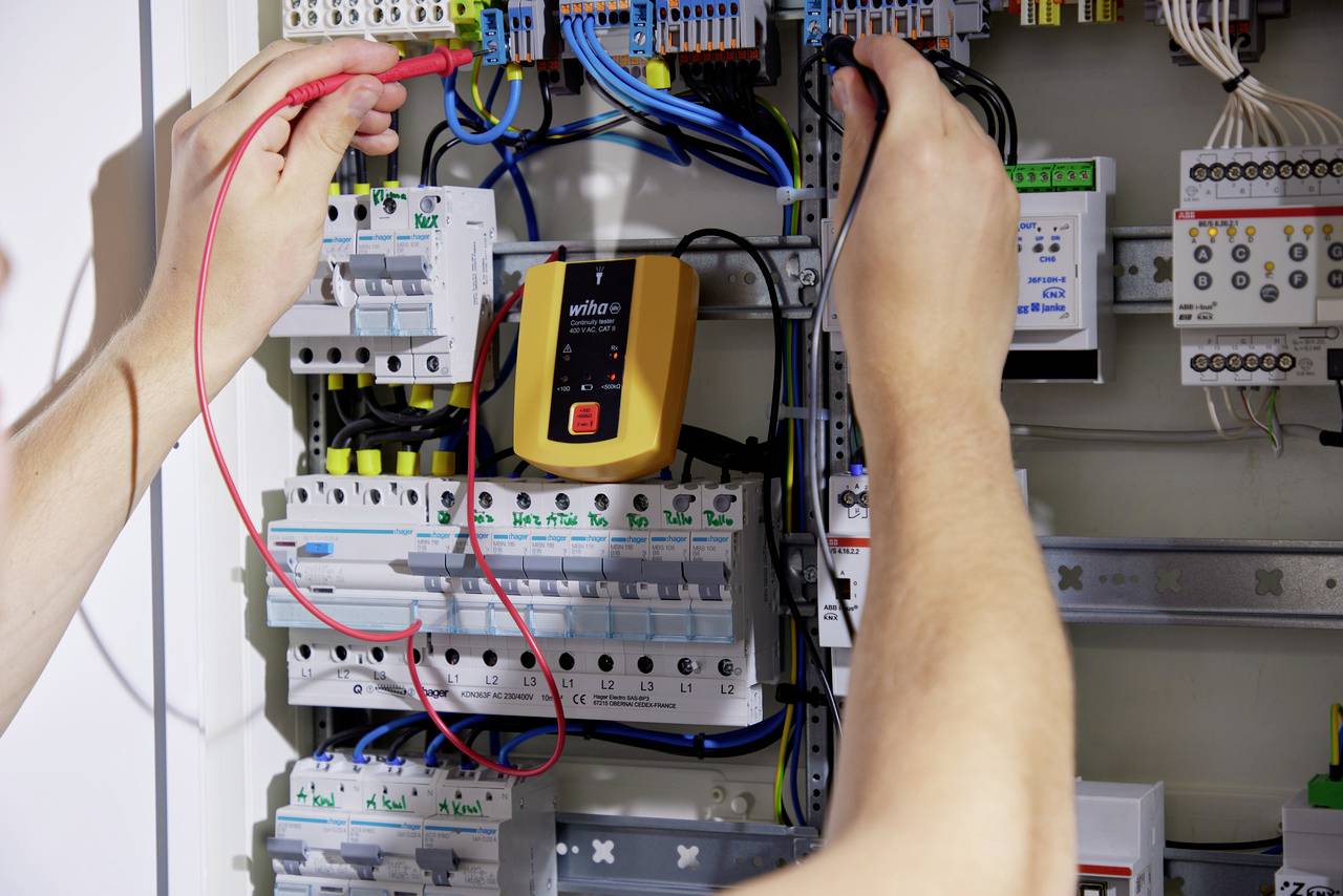 Een persoon test elektrische schakelingen in een zekeringenkast met een multimeter. Talrijke kleurrijke kabels zijn zichtbaar.