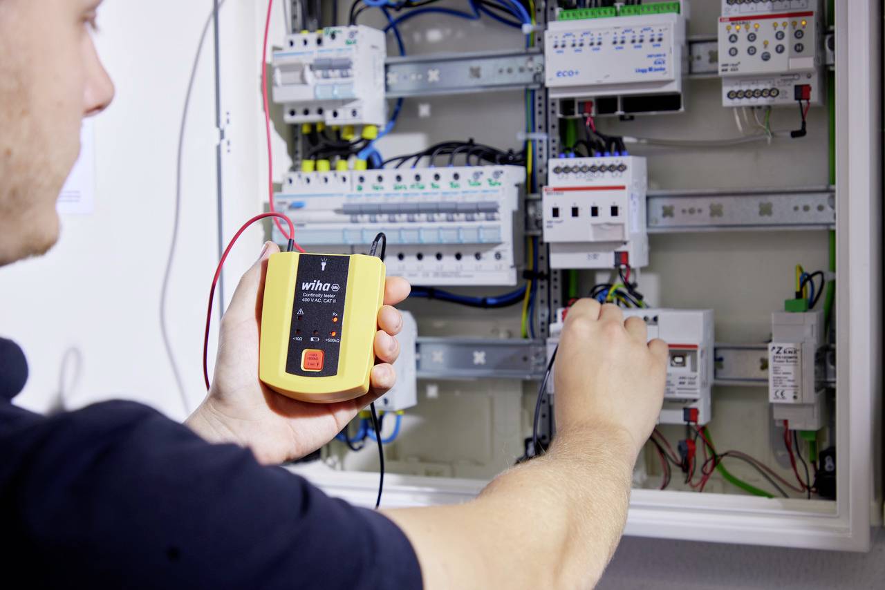 Een elektricien voert met een meettoestel een meting uit in een open schakelkast. Op de achtergrond zijn bekabeling en verschillende schakelapparatuur zichtbaar.