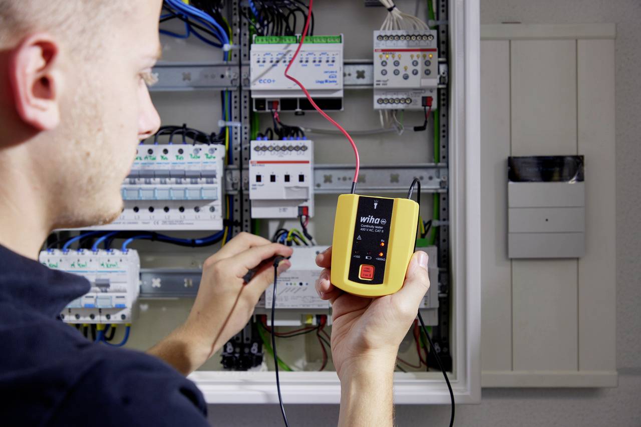 Een man test een elektrisch schakelbord met een spanningzoeker. Kabels en zekeringen zijn zichtbaar.