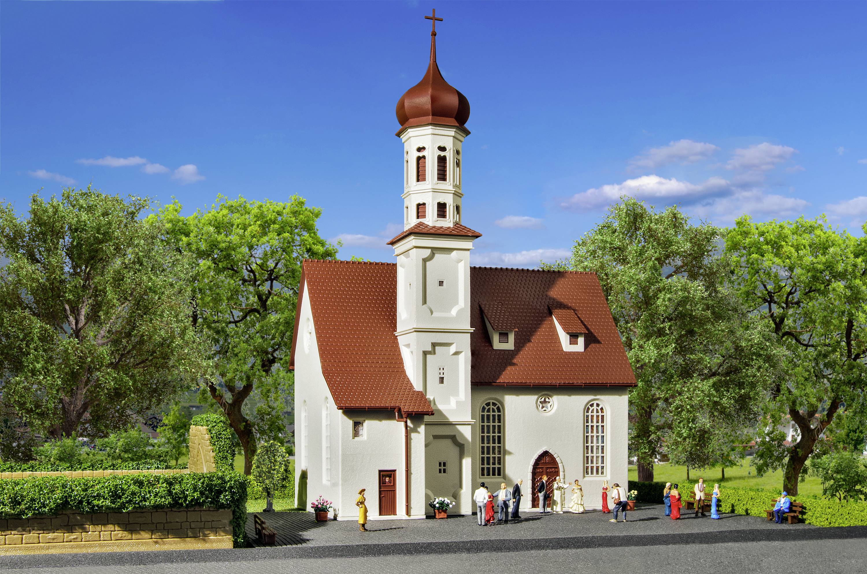Een kleine witte kerk met een rood dak en torenspits, omringd door bomen. Mensen staan voor de ingang.