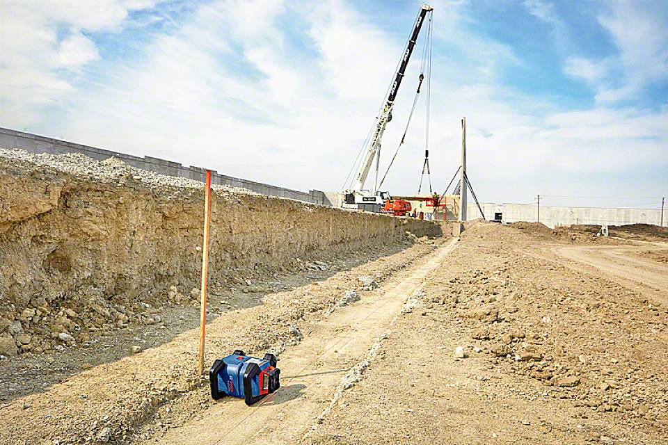 Een bouwplaats met een kraan die een grote betonplaat optilt. Op de voorgrond ligt een meetinstrument op de grond. Op de achtergrond is een gedeeltelijk opgebouwd bouwwerk te zien.