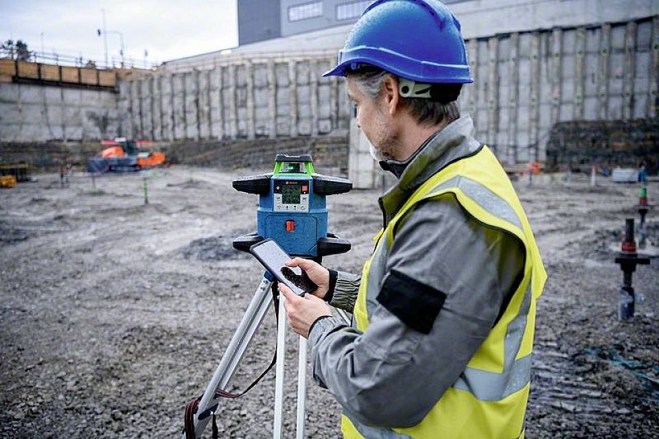 Een bouwvakker met helm en veiligheidsjas gebruikt een waterpas en een smartphone op een bouwterrein.