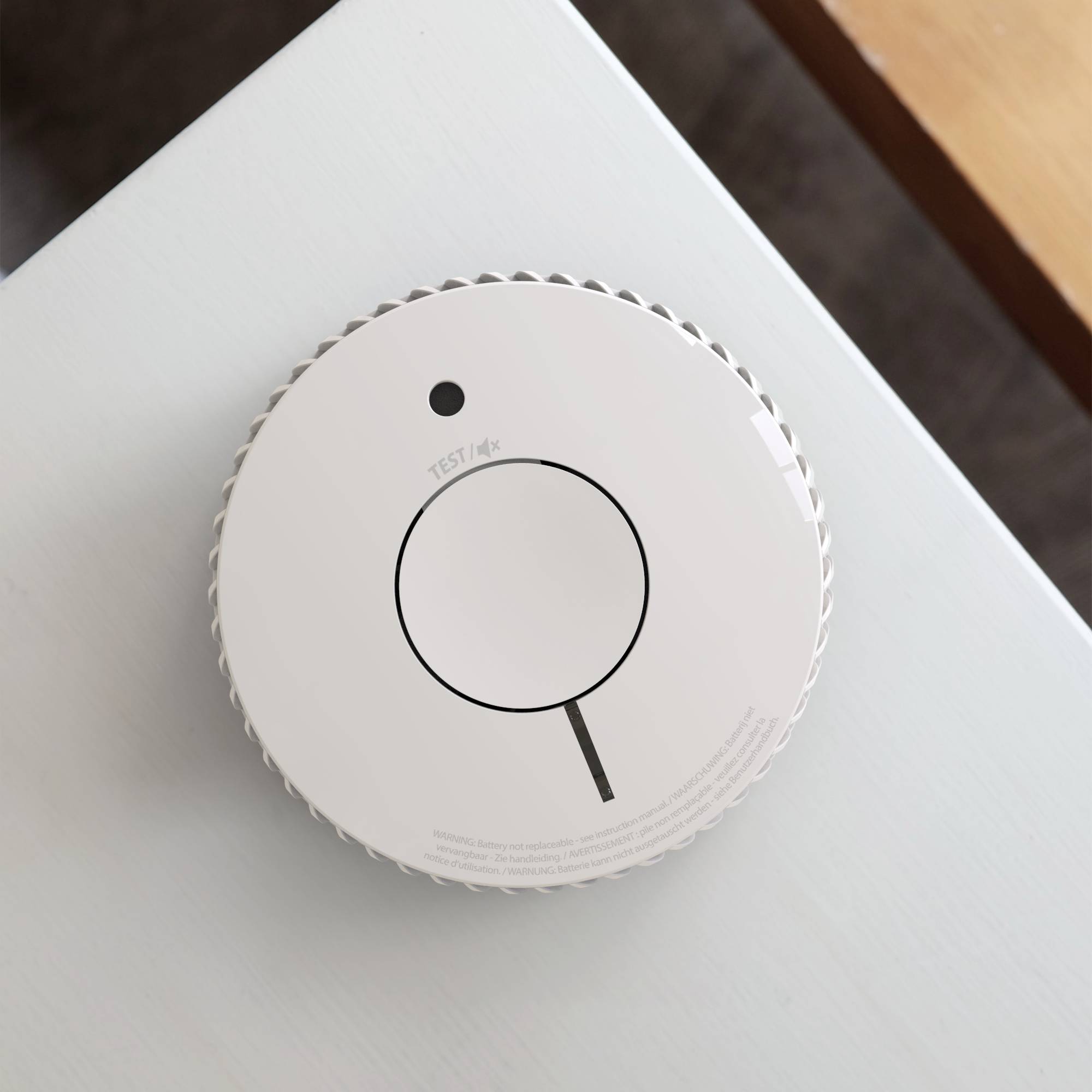 Een witte rookmelder is gemonteerd aan het plafond van een kamer. Aan de voorkant bevindt zich een testknop met het opschrift 'TEST'.