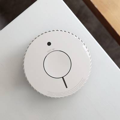 Een witte rookmelder is gemonteerd aan het plafond van een kamer. Aan de voorkant bevindt zich een testknop met het opschrift 'TEST'.