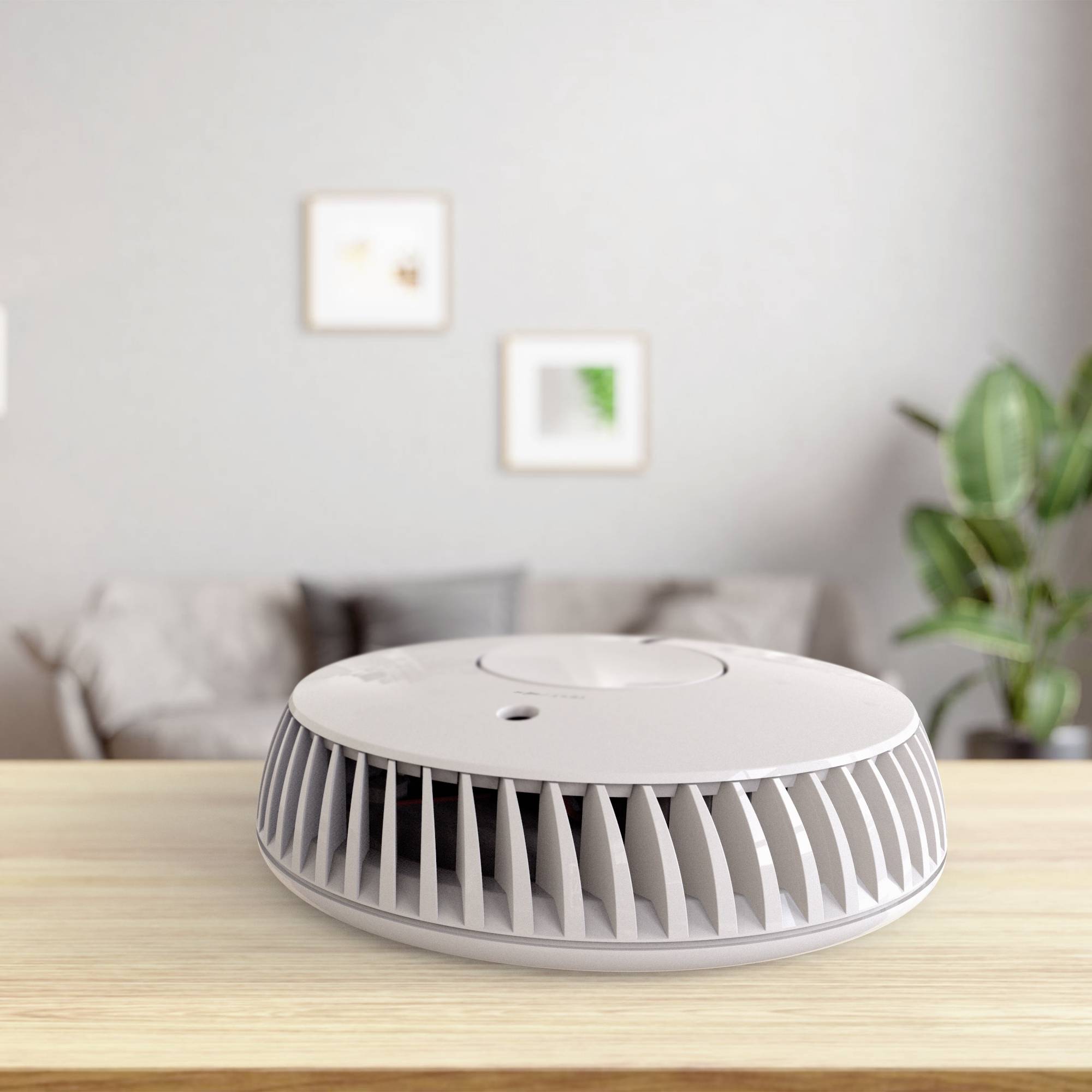 Een moderne rookmelder staat op een houten tafel in een woonkamer. Op de achtergrond zijn een bank en ingelijste afbeeldingen zichtbaar.
