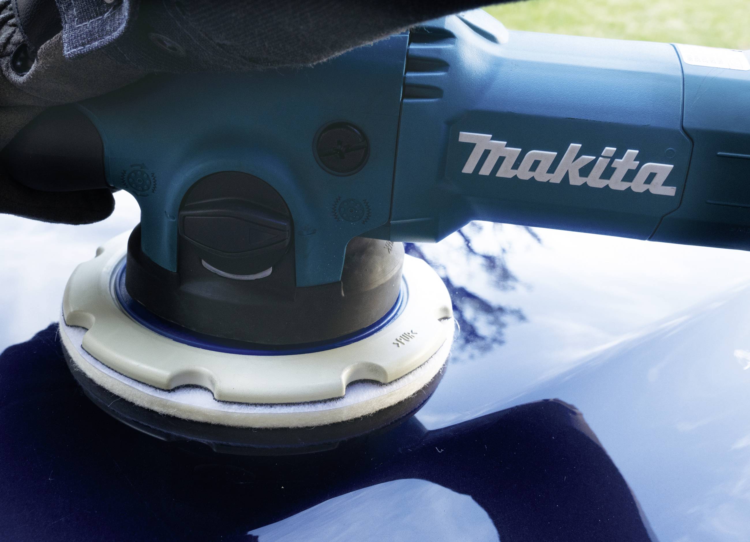 'Makita' polijstmachine polijst een glanzend blauwe oppervlakte. Handschoen houdt de machine vast. Buitengebied op de achtergrond.