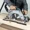 Een ambachtsman zaagt met een elektrische handcirkelzaag door een houten plank. De zaag produceert houtspaanders en stof.