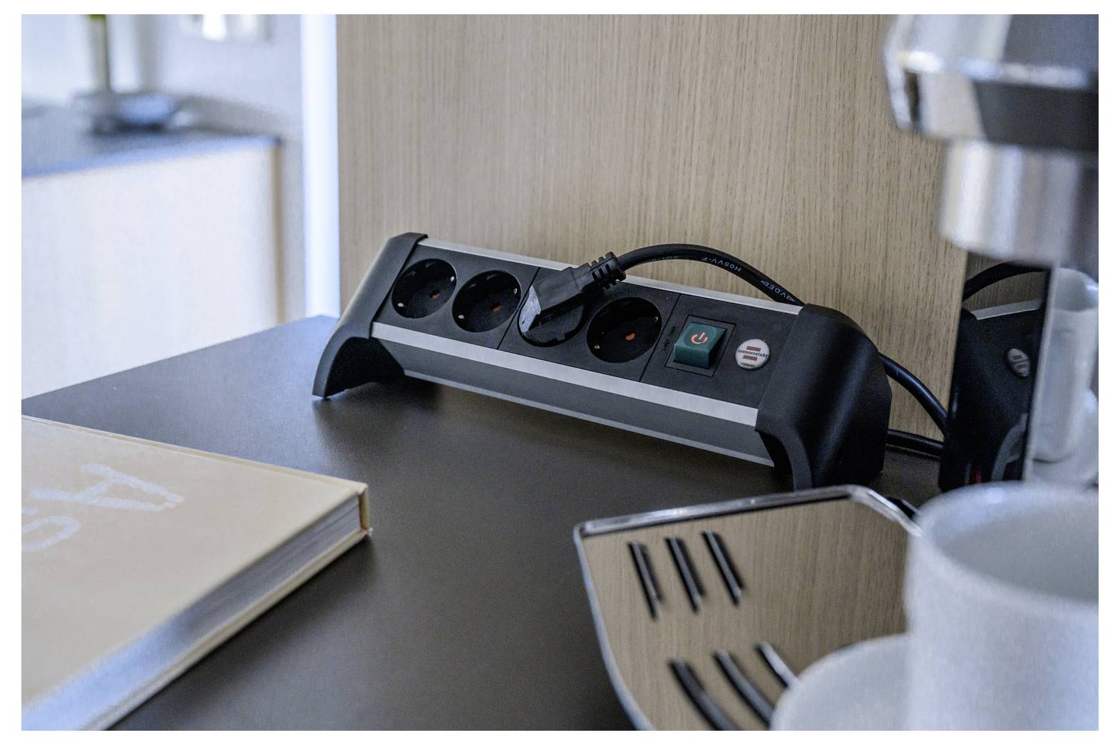 Een bureaubladstekkerstrip met meerdere outlets en een ingestalde stekker staat naast een boek en een koffiezetapparaat, wat een hotelwerkruimte suggereert.
