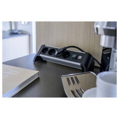 Een bureaubladstekkerstrip met meerdere outlets en een ingestalde stekker staat naast een boek en een koffiezetapparaat, wat een hotelwerkruimte suggereert.