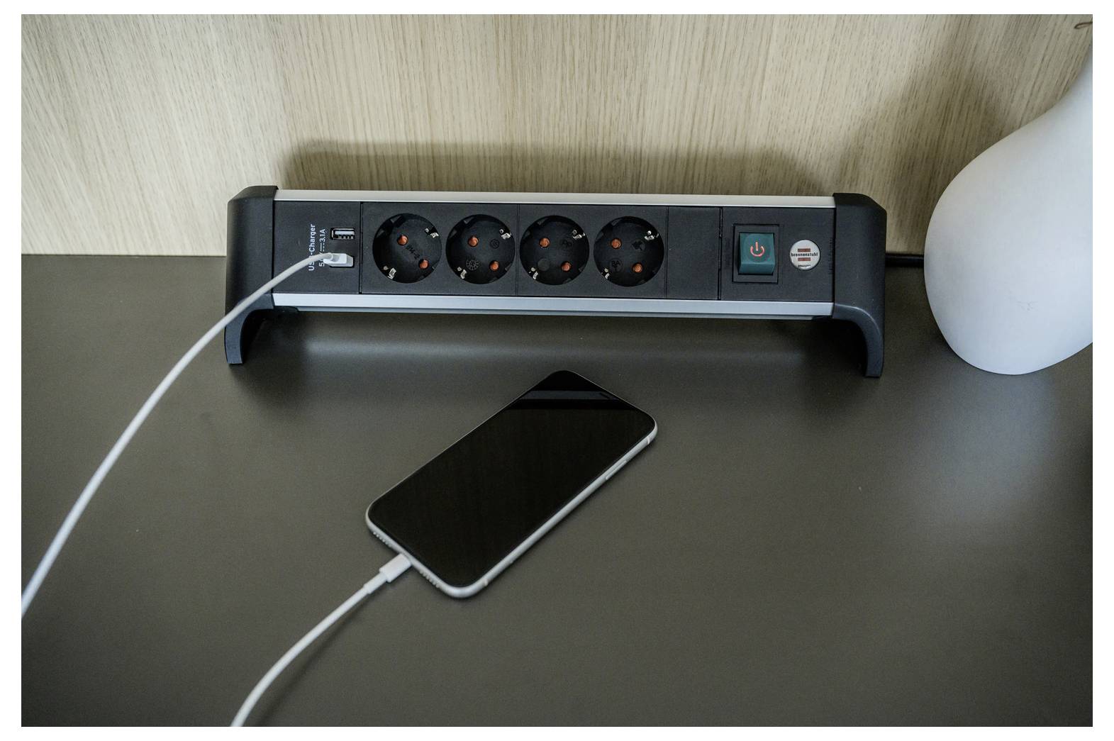 Een smartphone die op een bureau oplaadt naast een stekkerdoos met meerdere stopcontacten en een lamp.