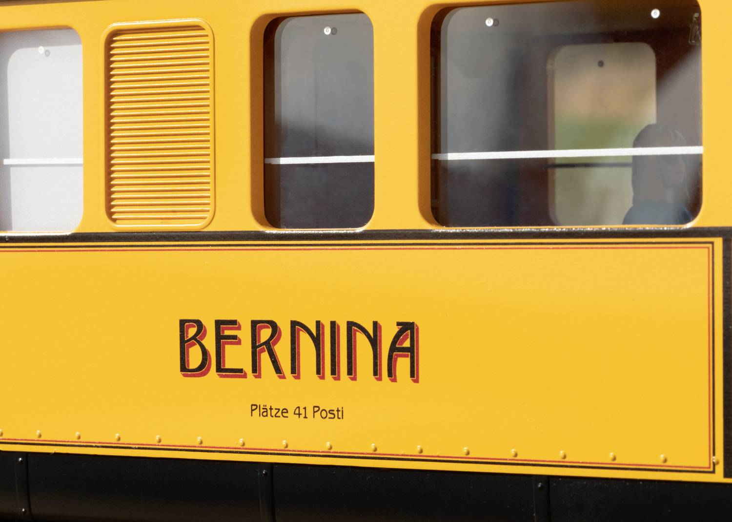 Een geel treincompartiment met het opschrift 'Bernina'. Daaronder staat 'Plaatsen 41 Post'. Ramen en details van de wagenwand zijn zichtbaar.