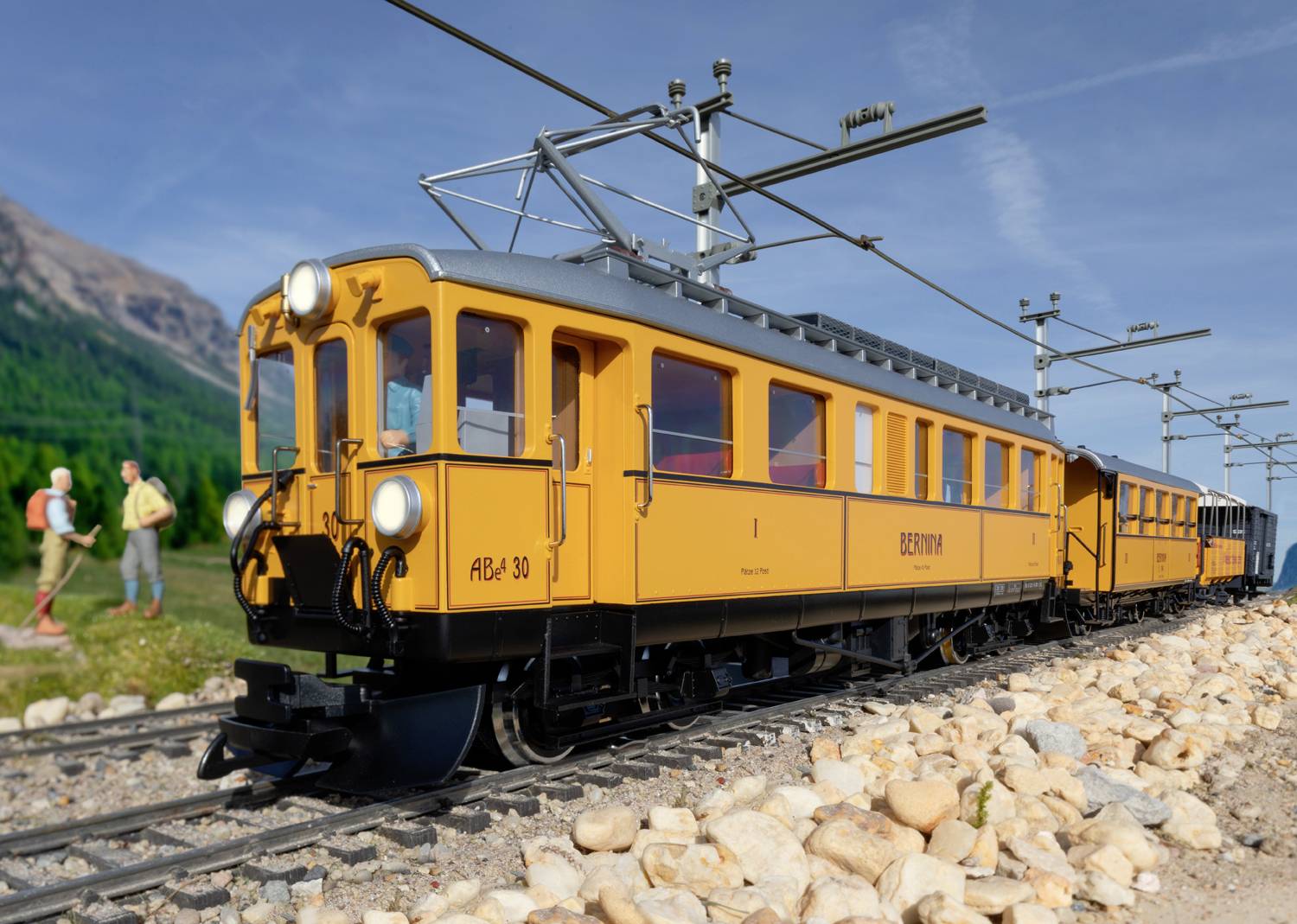 Gele trein rijdt op een smalspoorspoor door een landelijk landschap, met bergen op de achtergrond en twee personen langs de spoorbaan.