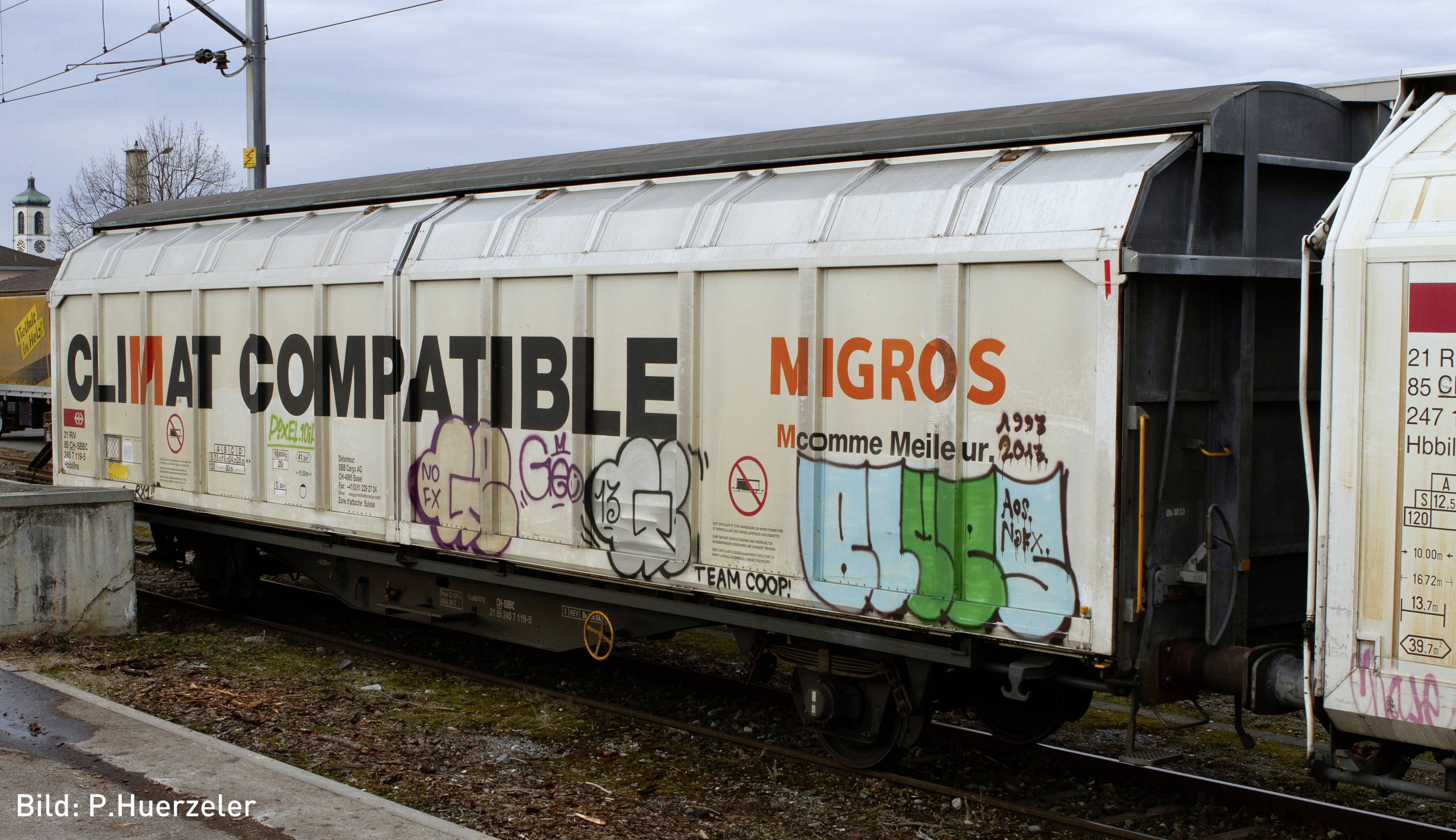 Goederenwagon met het opschrift 'CLIMAT COMPATIBLE' en graffiti. Op de achtergrond zijn spoorweginfrastructuur en een kerktoren zichtbaar.