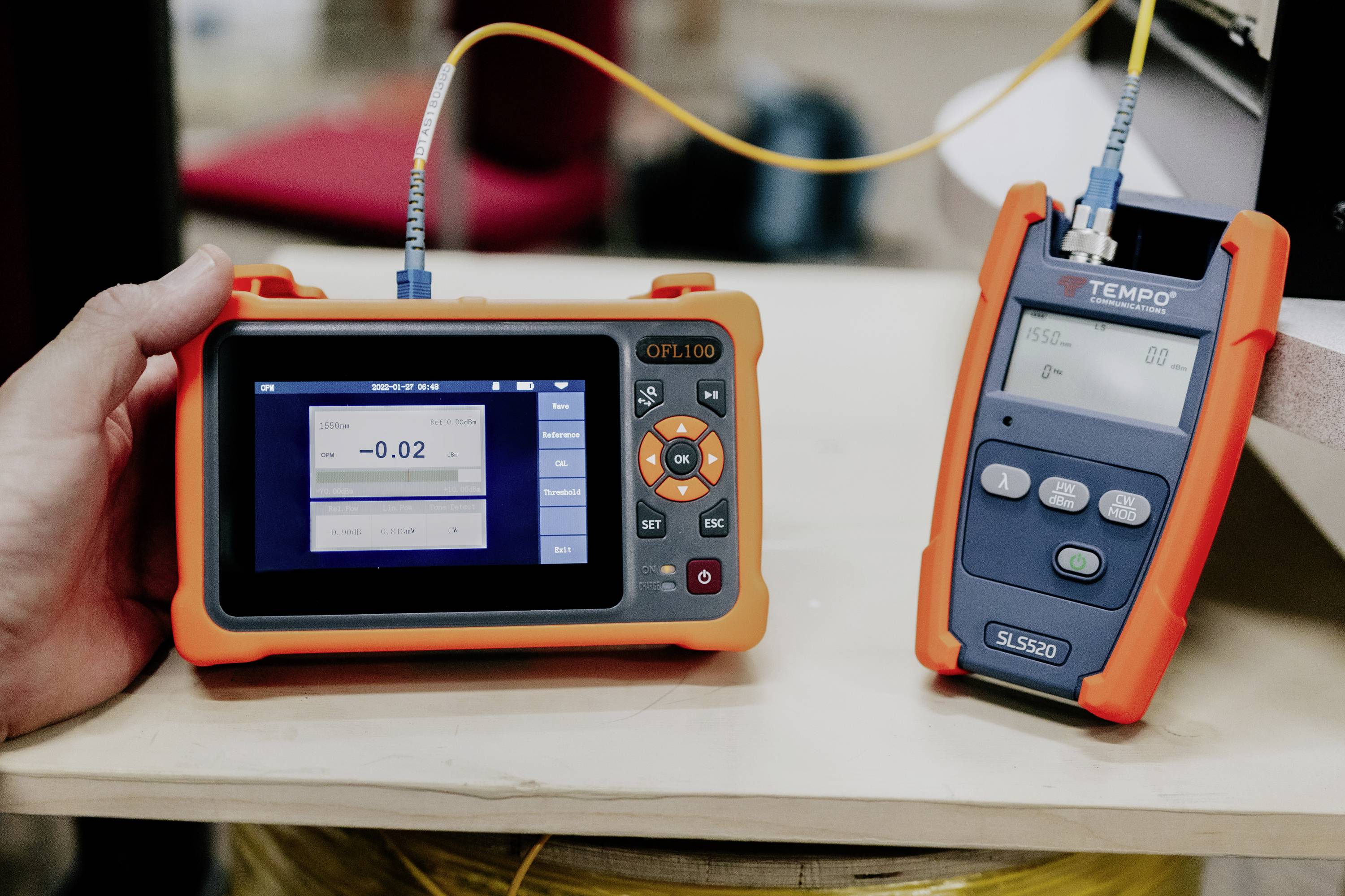 Een persoon houdt een handapparaat voor glasvezelcontrole met een display waarop '-0,02' wordt weergegeven. Naast het apparaat ligt nog een ander meetinstrument op een tafel.