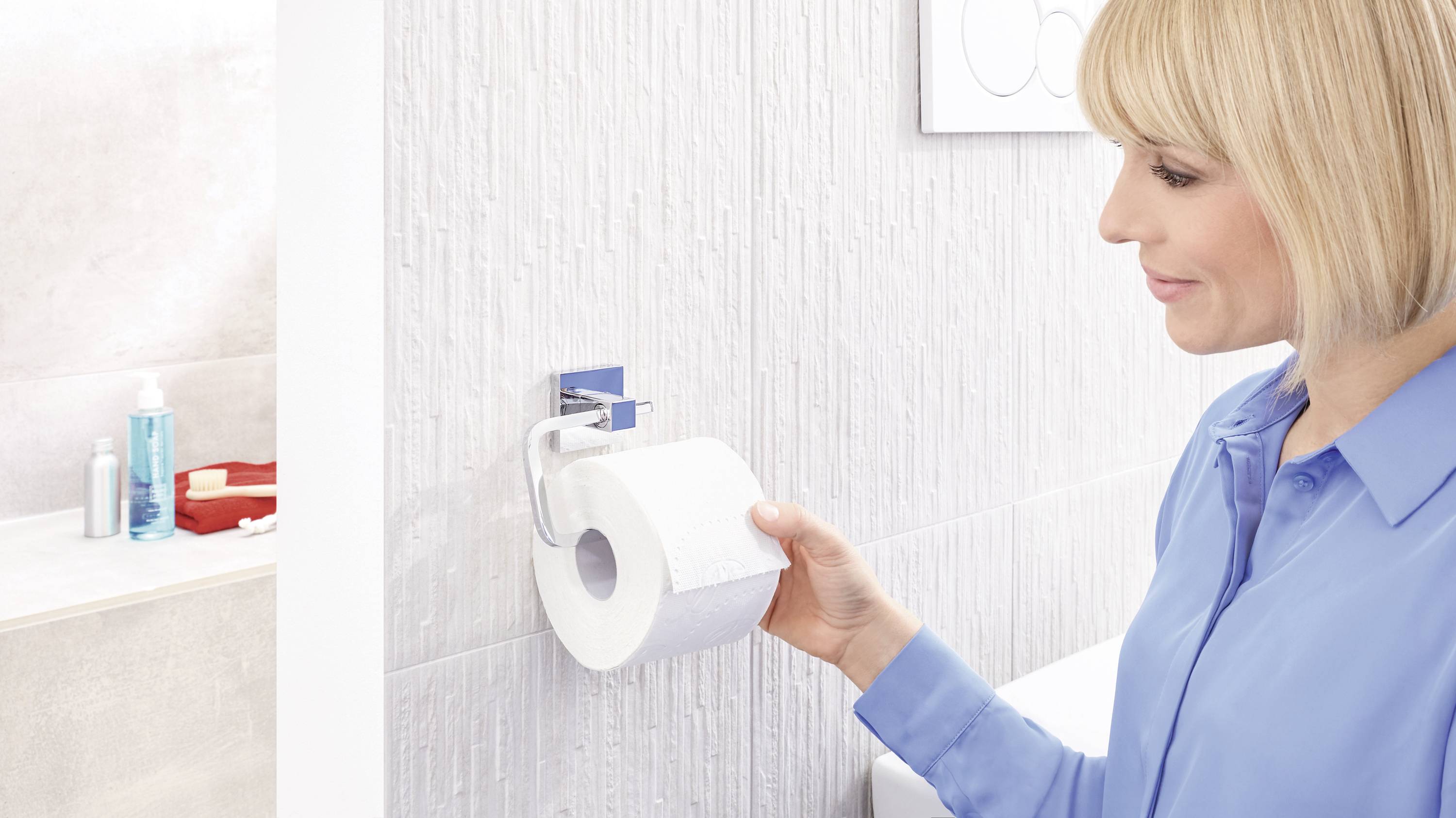 Een persoon in een blauw shirt verwisselt een toiletpapierrol in een lichte badkamer. Op de achtergrond zijn verzorgingsproducten zichtbaar.