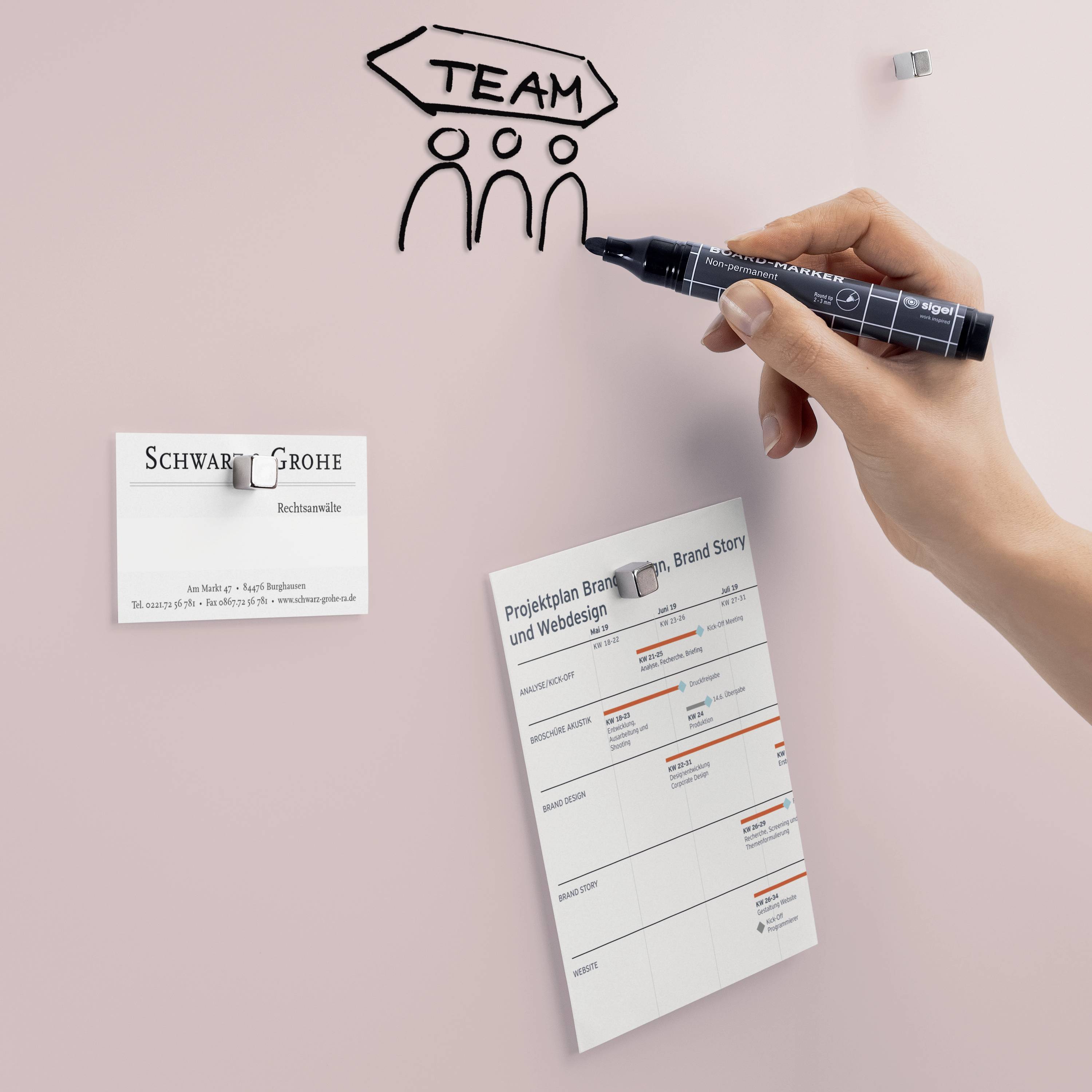 'Team' wordt met een zwarte pen op een roze muur geschilderd. Ernaast zijn een visitekaartje en een projectplanner bevestigd.