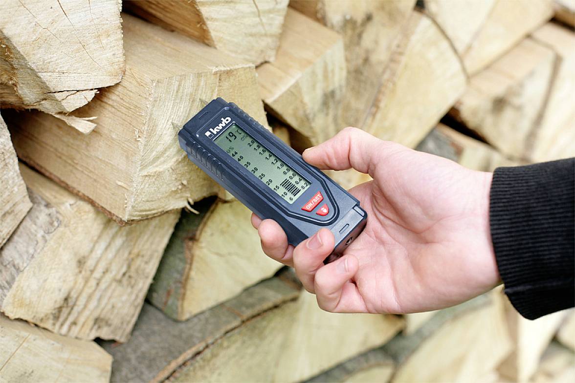 Een hand houdt een vochtigheidsmeter tegen een stapel gehakt brandhout om het vochtgehalte te meten.