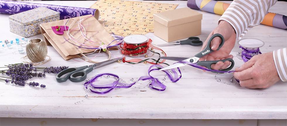 Een persoon knipt paars cadeaulint op een tafel die bedekt is met knutselmateriaal zoals een schaar, papier en andere decoratieve artikelen.