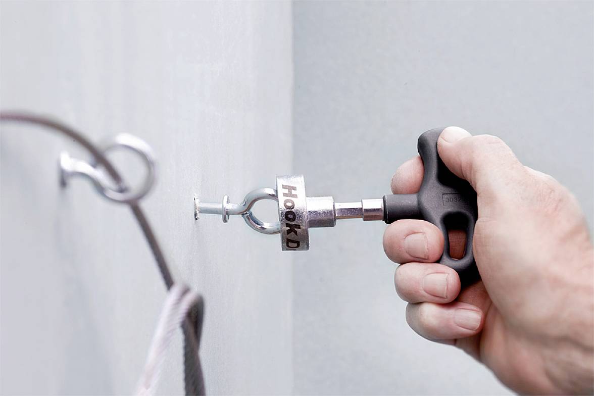 Een hand houdt een gereedschap vast om haken in een muur vast te zetten. Het gereedschap is gemarkeerd met 'HOOKD'.