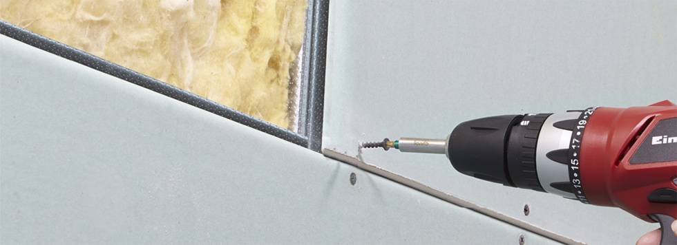 Een rode boormachine wordt gebruikt om een schroef in een blauwe gipsplaat te draaien. Op de achtergrond is isolatiemateriaal zichtbaar.