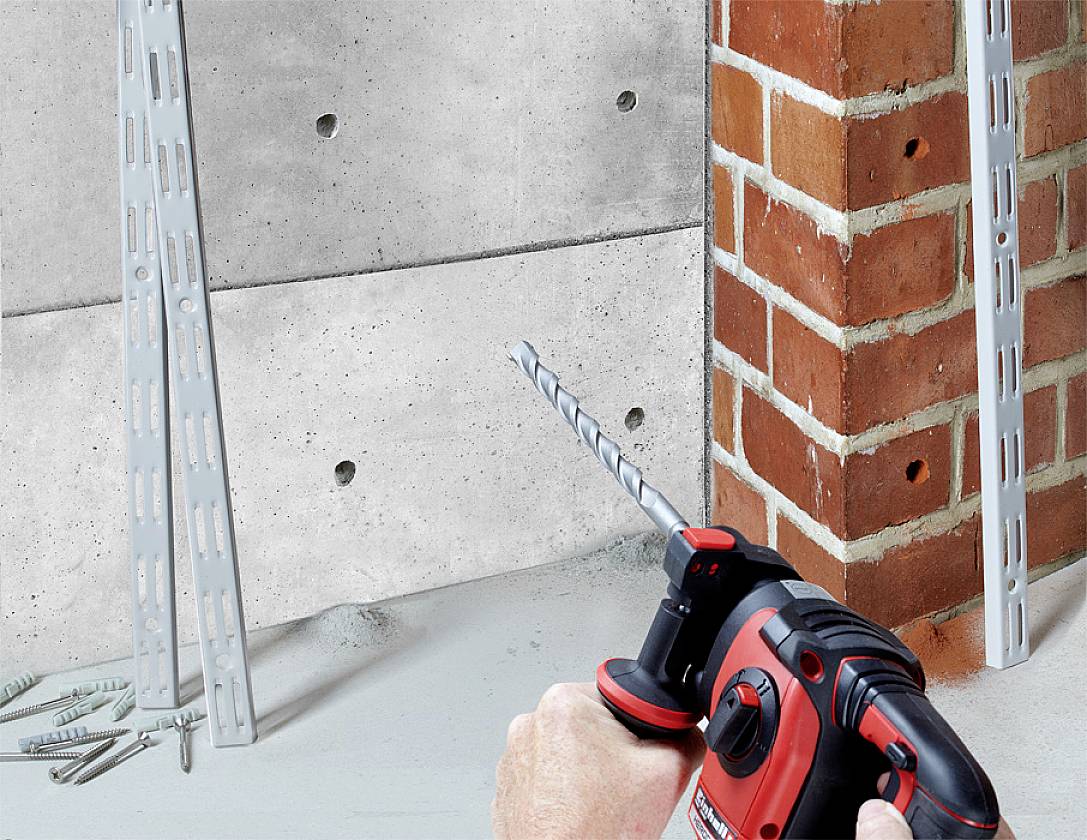 Een persoon houdt een boormachine en boort in een bakstenen muur. Op de achtergrond zijn beton- en bakstenen muren met bevestigingen zichtbaar.