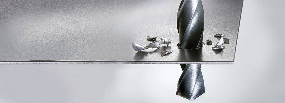 Een metalen boor doorboort een dun metalen plaatje, terwijl metaalspanen verspreid liggen rond het geboorde gat.