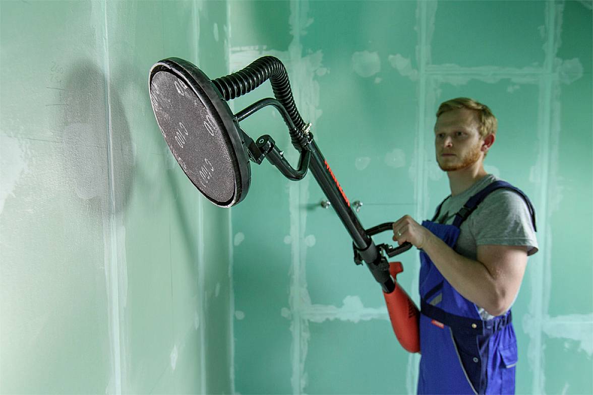 Een man slijpt een muur in een kamer met een slijpmachine. Hij draagt blauwe werkkleding. De muur heeft een groene tint.