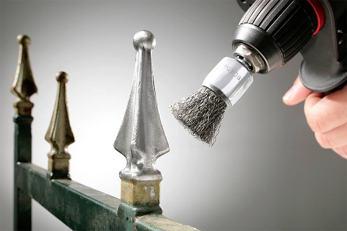 Een hand gebruikt een boormachine met een staalborstelopzetstuk om roest van een metalen hekpaal te verwijderen.