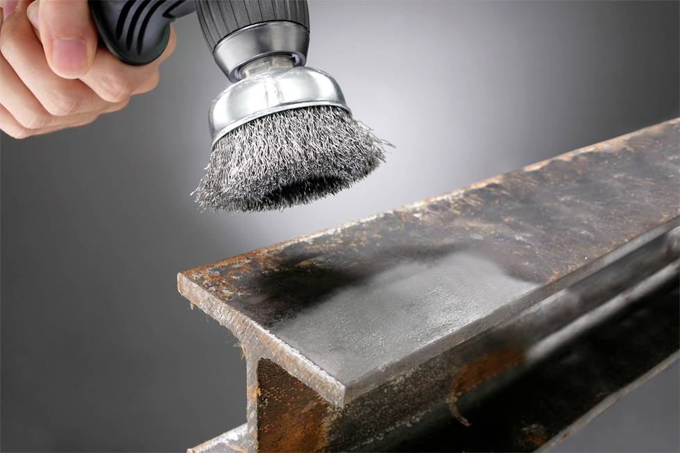 Een hand houdt een draadborstel vast, waarbij een roestige metalen balk wordt gepolijst, waardoor een gladde, glanzende oppervlakte zichtbaar wordt. De achtergrond is een neutrale grijstint.