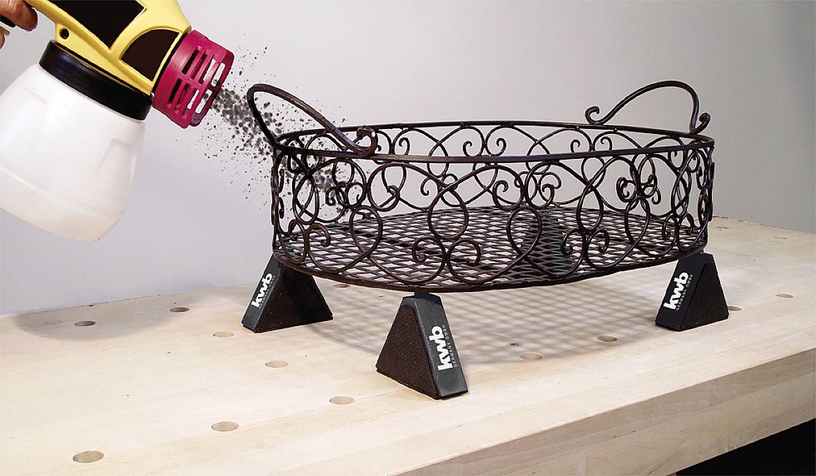 'Een metalen mand wordt op een tafel met een spuitpistool zwart gespoten. Kleine, zwarte lakdruppels verdelen zich gelijkmatig.'