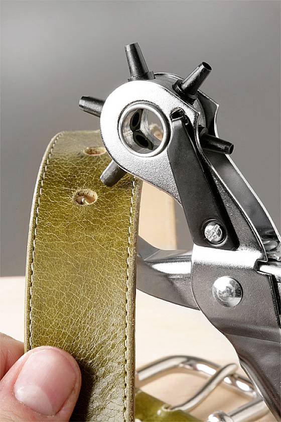 Een persoon ponst gaten in een olijfgroene leren riem met een perforator op een lichte achtergrond.